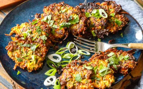 Knusprige Zucchini Kartoffel Puffer, garniert mit frischen Kräutern und Frühlingszwiebeln, liegen auf einem blauen Teller. Eine Gabel liegt daneben.