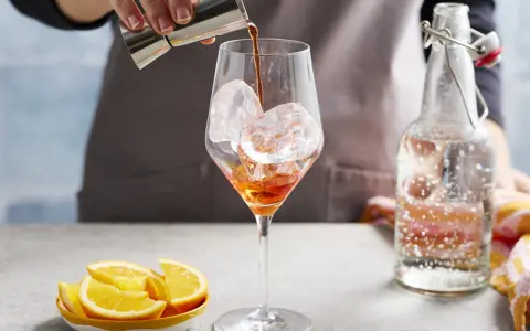 Zabawne przygotowanie Aperol Spritz Rezept Original. Likier wlewa się do kieliszka z lodem. Obok leżą pomarańcze i butelka prosecco.