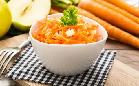 Ein frischer Karotten-Apfel-Salat, garniert mit Petersilie, steht auf einem karierten Tuch. Im Hintergrund liegen ganze Karotten und ein halber Apfel.