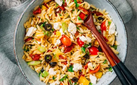 Bunter Kritharaki Salat mit Tomaten, Oliven, Feta und Kräutern, serviert in einer Schüssel mit Holzstäbchen.