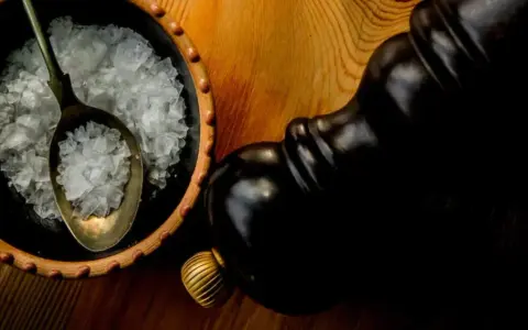 Fleur de sel, zarten Salzkristallen auf einem Löffel in einer Schale, neben einer dunklen Pfeffermühle.