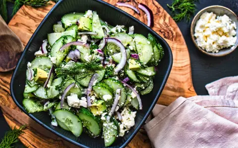 Erfrischender gurken feta salat mit Avocado, roten Zwiebeln und Dill, serviert in einer schwarzen Schüssel auf einem Holzbrett.