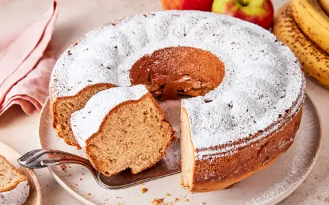 Saftiger Apfel-Bananen-Kuchen, bestäubt mit Puderzucker. Daneben liegen frische Äpfel und Bananen.