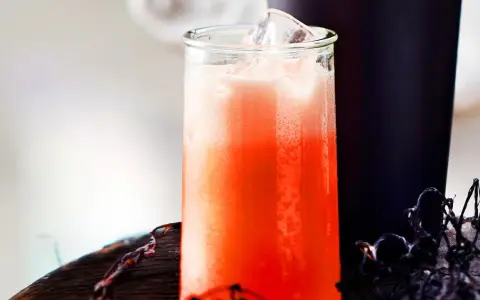 Erfrischender Aperol Sour mit Eiswürfeln in einem Glas, daneben ein dunkler Becher und Spinnweben-Deko.