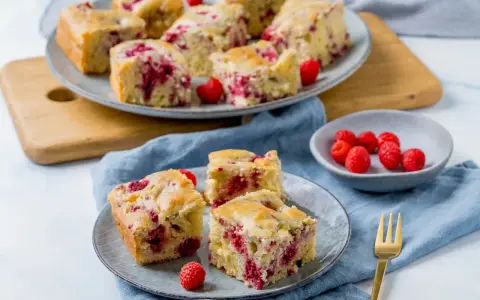 Saftiger Kuchen mit Himbeeren, auf Tellern und einer Platte angerichtet, daneben eine Schale mit frischen Himbeeren.