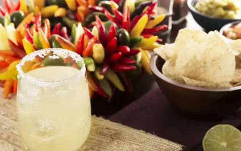 Ein erfrischendes Margarita-Rezept: Ein Glas mit Salzrand, Eiswürfeln und Limette, daneben bunte Chilis, Guacamole und Tortilla-Chips.