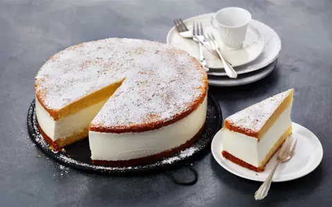 Eine köstliche Quark Sahne Torte, mit Puderzucker bestäubt, wartet auf ihre Genießer. Ein Stück ist bereits auf einem Teller angerichtet.