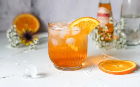 Erfrischender Crodino Spritz mit Eiswürfeln und Orangenscheibe, dekoriert mit kleinen weißen Blüten.