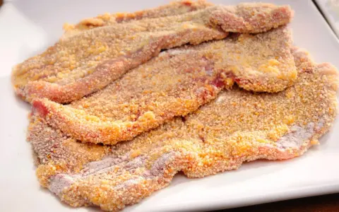 Mehrere rohe Schnitzel liegen auf einem weißen Teller, bereit zum Schnitzel panieren. Die Panade ist goldbraun und knusprig.