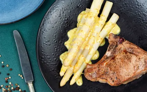 Ein saftiges Schweinekotelett liegt neben einem Haufen cremigen Spargel in der Pfanne, beträufelt mit einer würzigen Sauce.