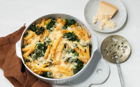 Herzhafter nudelauflauf mit spinat, überbacken mit Käse. Daneben ein Stück Parmesan und ein dekoratives Servierbesteck.