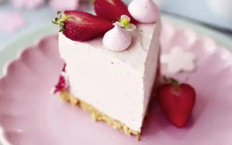 Ein Stück Erdbeer Cheesecake ohne Backen, dekoriert mit frischen Erdbeeren und rosa Baiser-Tupfern auf einem rosa Teller.