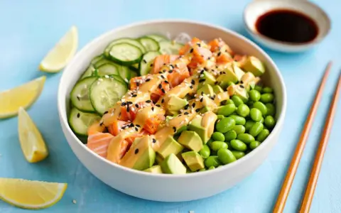 Schüssel mit einem frischen Salat aus Lachs, Avocado, Edamame und Gurkenscheiben, beträufelt mit cremiger Sauce und Sesam. Zitronenspalten und Sojasauce daneben.