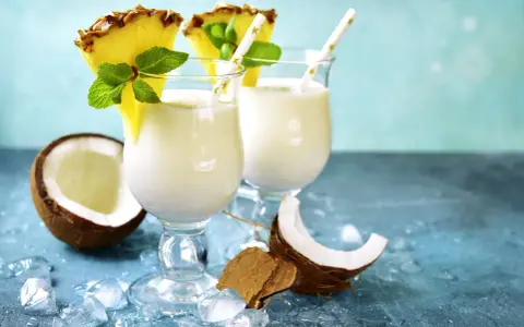Zwei cremige Malibu Cocktails mit Ananas und Minze, umgeben von Kokosnuss und Eis.