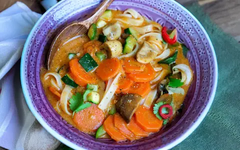 Eine dampfende, cremige Thai Curry Suppe mit Karotten, Zucchini, Pilzen und Reisnudeln in einer lila Schüssel.