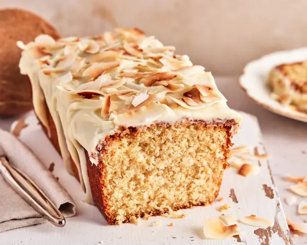 Saftiger Kuchen mit Kokosmilch, überzogen mit weißer Glasur und garniert mit gerösteten Kokoschips. Daneben eine Kokosnuss.