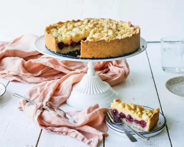 Ein saftiger Kirschkuchen mit Pudding und Streuseln auf einem Tortenständer. Ein Stück ist bereits auf einem Teller angerichtet.