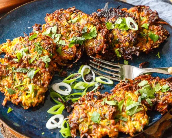 Knusprige Zucchini Kartoffel Puffer, garniert mit frischen Kräutern und Frühlingszwiebeln, liegen auf einem blauen Teller. Eine Gabel liegt daneben.