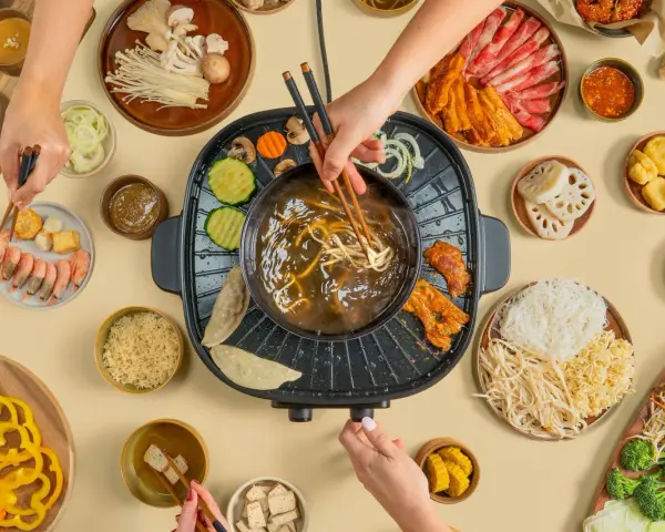 Hände greifen nach Zutaten für einen Hot Pot. Fleisch, Gemüse, Garnelen und Nudeln werden in die köchelnde Brühe getaucht.