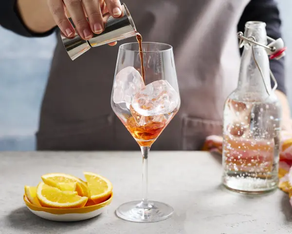 Zabawne przygotowanie Aperol Spritz Rezept Original. Likier wlewa się do kieliszka z lodem. Obok leżą pomarańcze i butelka prosecco.