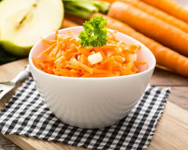 Ein frischer Karotten-Apfel-Salat, garniert mit Petersilie, steht auf einem karierten Tuch. Im Hintergrund liegen ganze Karotten und ein halber Apfel.