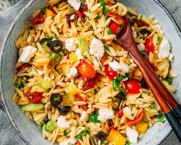 Bunter Kritharaki Salat mit Tomaten, Oliven, Feta und Kräutern, serviert in einer Schüssel mit Holzstäbchen.