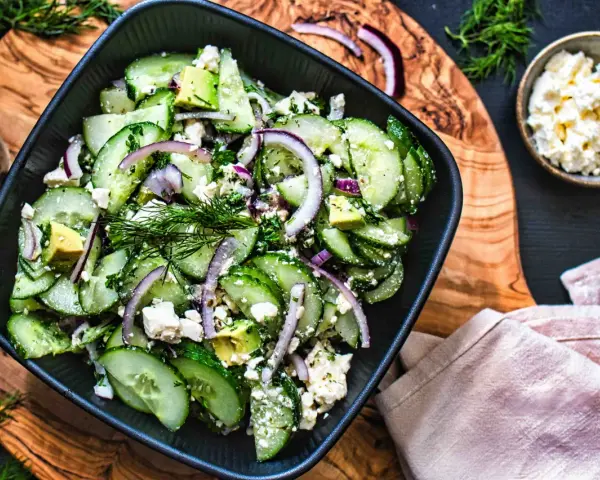 Erfrischender gurken feta salat mit Avocado, roten Zwiebeln und Dill, serviert in einer schwarzen Schüssel auf einem Holzbrett.