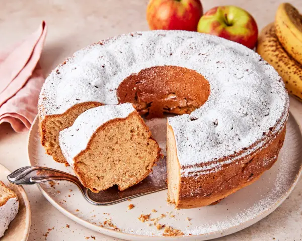 Saftiger Apfel-Bananen-Kuchen, bestäubt mit Puderzucker. Daneben liegen frische Äpfel und Bananen.