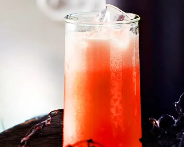 Erfrischender Aperol Sour mit Eiswürfeln in einem Glas, daneben ein dunkler Becher und Spinnweben-Deko.