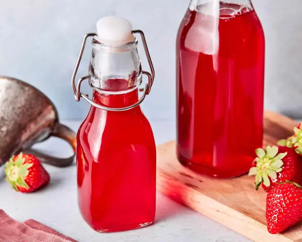 Zwei Flaschen mit leuchtend rotem Erdbeersirup, frisch zubereitet. Frische Erdbeeren liegen daneben.
