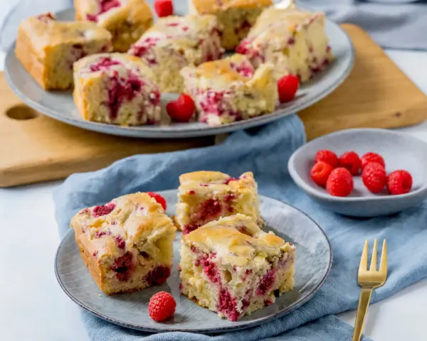 Saftiger Kuchen mit Himbeeren, auf Tellern und einer Platte angerichtet, daneben eine Schale mit frischen Himbeeren.