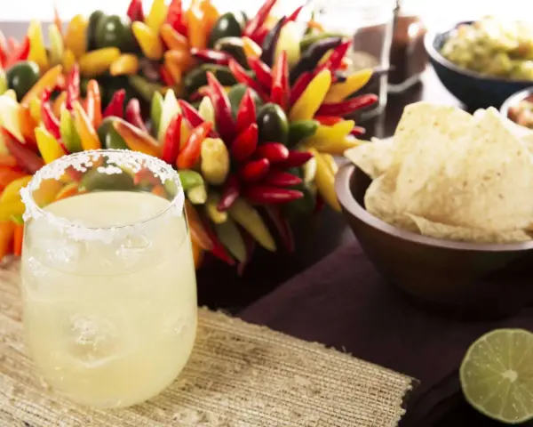 Ein erfrischendes Margarita-Rezept: Ein Glas mit Salzrand, Eiswürfeln und Limette, daneben bunte Chilis, Guacamole und Tortilla-Chips.