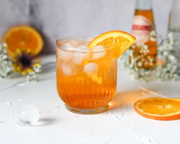Erfrischender Crodino Spritz mit Eiswürfeln und Orangenscheibe, dekoriert mit kleinen weißen Blüten.