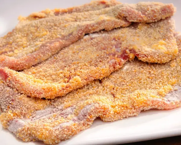 Mehrere rohe Schnitzel liegen auf einem weißen Teller, bereit zum Schnitzel panieren. Die Panade ist goldbraun und knusprig.