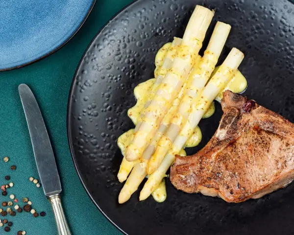 Ein saftiges Schweinekotelett liegt neben einem Haufen cremigen Spargel in der Pfanne, beträufelt mit einer würzigen Sauce.