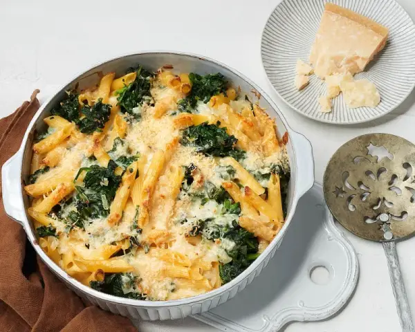 Herzhafter nudelauflauf mit spinat, überbacken mit Käse. Daneben ein Stück Parmesan und ein dekoratives Servierbesteck.