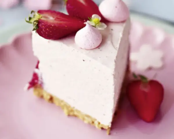 Ein Stück Erdbeer Cheesecake ohne Backen, dekoriert mit frischen Erdbeeren und rosa Baiser-Tupfern auf einem rosa Teller.