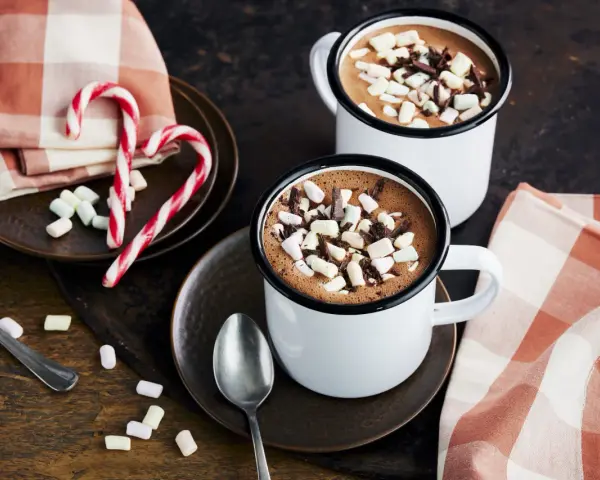 Zwei Tassen heißer Kakao mit Marshmallows und Schokostückchen, daneben Zuckerstangen und eine karierte Serviette.