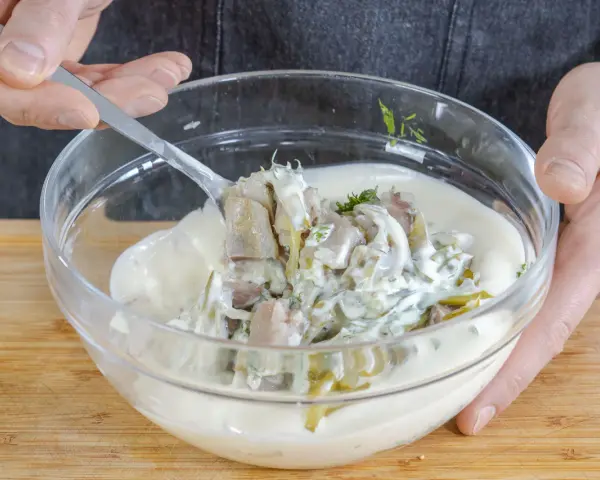 Matjes Hausfrauenart, Omas Rezept: Ein Löffel voll cremiger Sauce mit Matjesstücken und Dill wird aus einer Glasschüssel gehoben.
