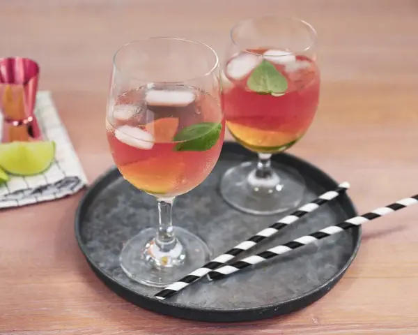 Zwei Gläser Rosato Spritz mit Eis und Minze, dazu Limettenspalten und ein Messbecher. Perfekt für einen sommerlichen Genuss.