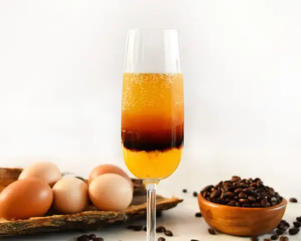Schicht-Cocktail mit Ei, Kaffee und Orangensaft. Daneben liegen Eier und Kaffeebohnen.