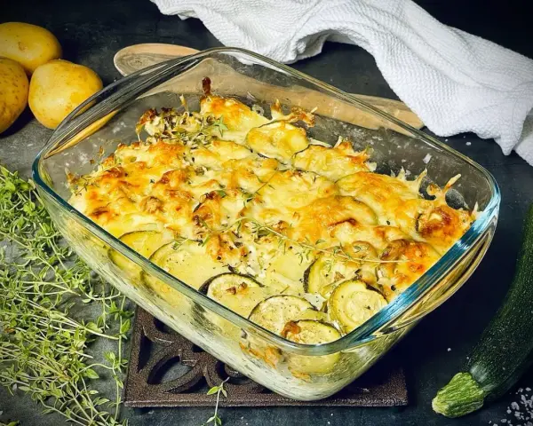 Goldbrauner Zucchini-Kartoffel-Auflauf mit Käse überbacken, garniert mit frischem Thymian. Daneben liegen Kartoffeln und eine Zucchini.