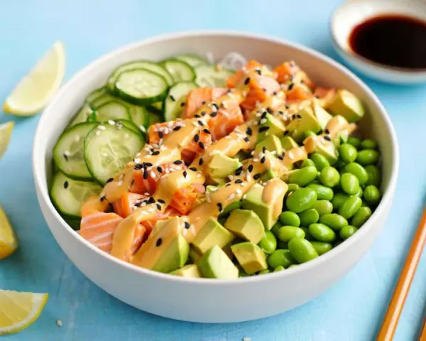 Schüssel mit einem frischen Salat aus Lachs, Avocado, Edamame und Gurkenscheiben, beträufelt mit cremiger Sauce und Sesam. Zitronenspalten und Sojasauce daneben.