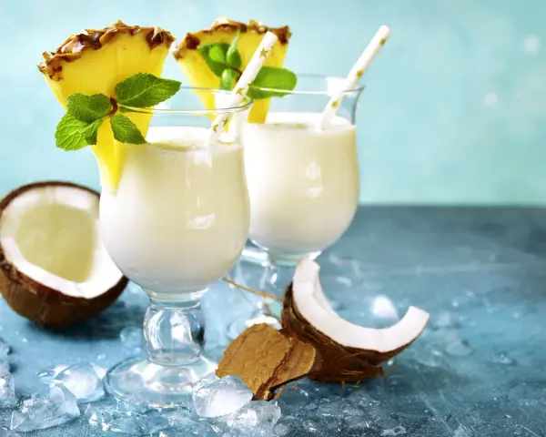 Zwei cremige Malibu Cocktails mit Ananas und Minze, umgeben von Kokosnuss und Eis.