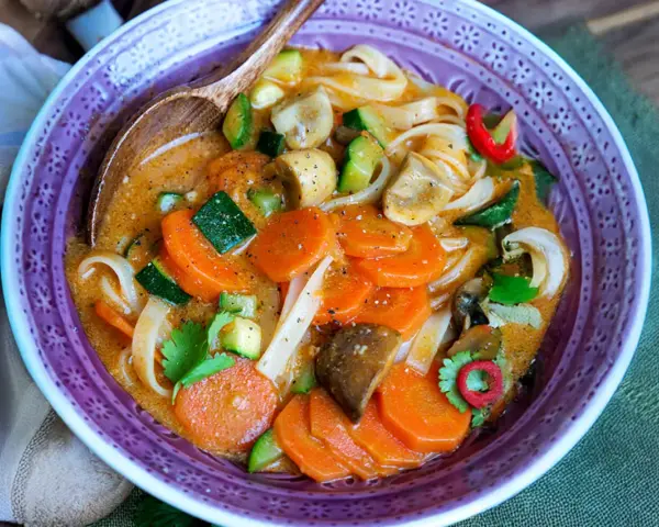 Eine dampfende, cremige Thai Curry Suppe mit Karotten, Zucchini, Pilzen und Reisnudeln in einer lila Schüssel.