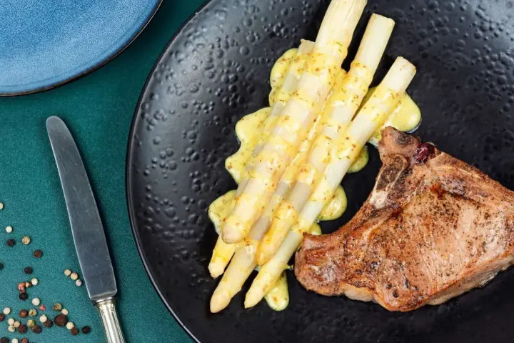 Ein saftiges Schweinekotelett liegt neben einem Haufen cremigen Spargel in der Pfanne, beträufelt mit einer würzigen Sauce.