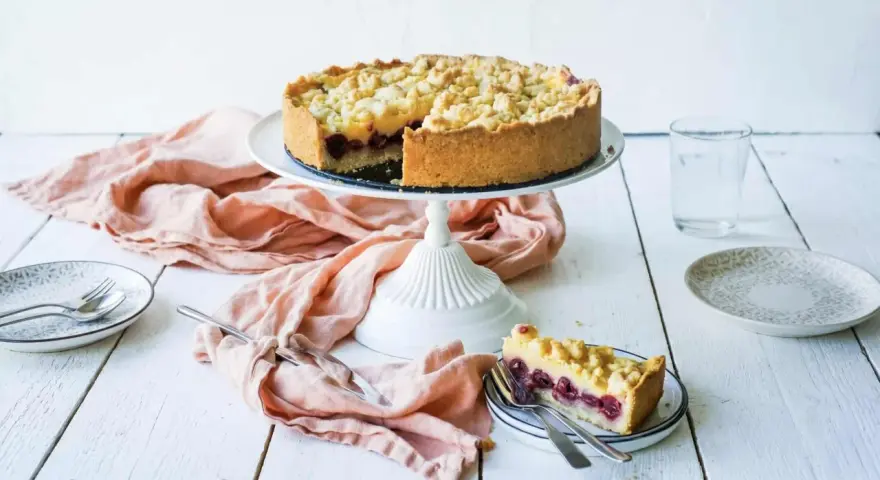 Ein saftiger Kirschkuchen mit Pudding und Streuseln auf einem Tortenständer. Ein Stück ist bereits auf einem Teller angerichtet.