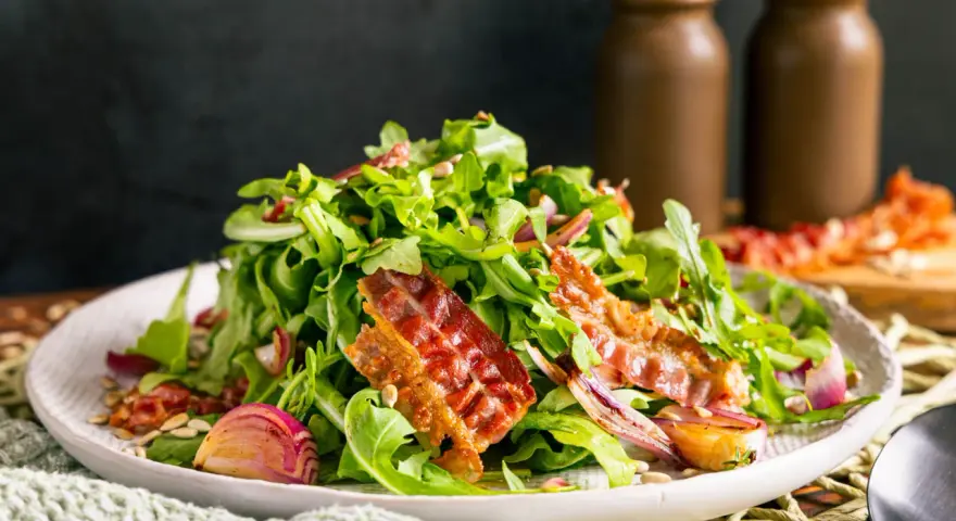 Warmer Rucolasalat mit knusprigem Speck, gerösteten Zwiebeln und Sonnenblumenkernen. Ein köstliches Gericht für Rucola Rezepte warm.