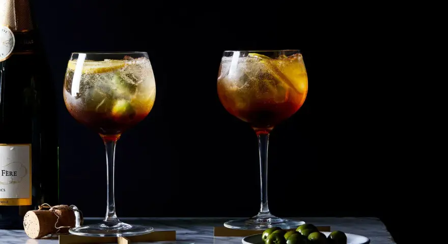 Zwei erfrischende Cynar Spritz Cocktails mit Eis, Zitrone und Oliven, daneben eine Champagnerflasche.