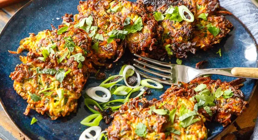 Knusprige Zucchini Kartoffel Puffer, garniert mit frischen Kräutern und Frühlingszwiebeln, liegen auf einem blauen Teller. Eine Gabel liegt daneben.